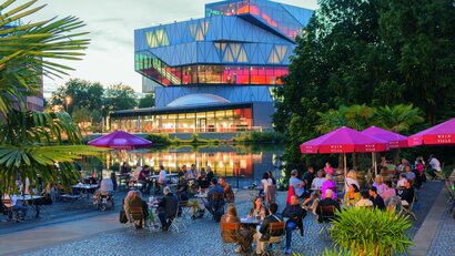 Das Bild zeigt den viel genutzten Außenbereich eines Lokals an einem Sommerabend. Im Freien stehen diverse Tische, Sonnenschirme und Pflanzen. Im Hintergrund sieht man einen See und ein großes Gebäude, das bunt beleuchtet ist. 
