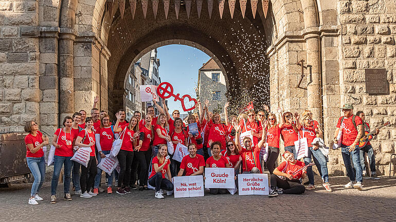 Bei dem Bild handelt es sich um ein Gruppenfoto mit 34 Personen, die alle ein rotes T-Shirt tragen. Manche halten beschriftete Schilder in der Hand oder schmeißen Konfetti in die Luft.