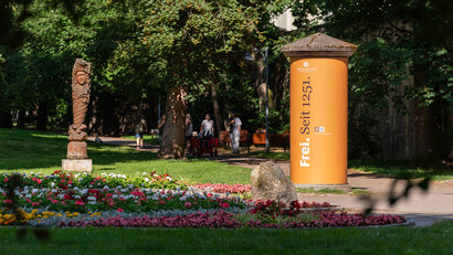 Das Foto zeigt einen Ausschnitt aus einem Park mit Blumenbepflanzung, einer Statue und Sitzmöglichkeiten.