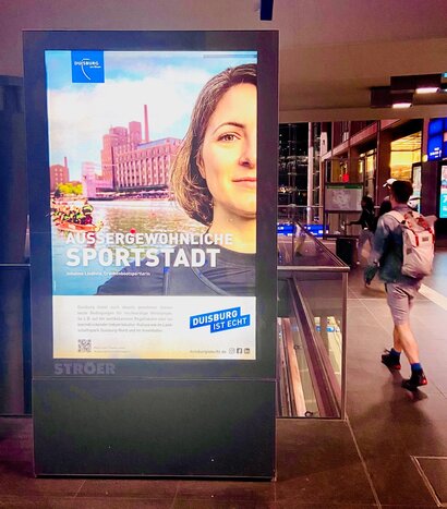 Das Foto ist aufgenommen in einem Bahnhof und zeigt eine Werbetafel. Auf dieser ist ein Bild von Ruderern auf dem Wasser sowie eine einzelne Ruderin im Vordergrund zu sehen. Es handelt sich hierbei um eine Anzeige, die Duisburg als "außergewöhnliche Sportstadt" bewirbt.
