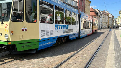 Auf dem Foto sieht man einen Fußgängerweg und eine Fahrbahn mit Schienen sowie eine Straßenbahn mit drei Waggons.
