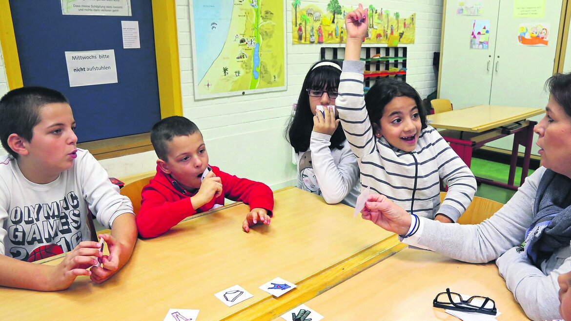 Klassenzimmer, Kinder sitzen an einem Tisch und ein Mädchen meldet sich