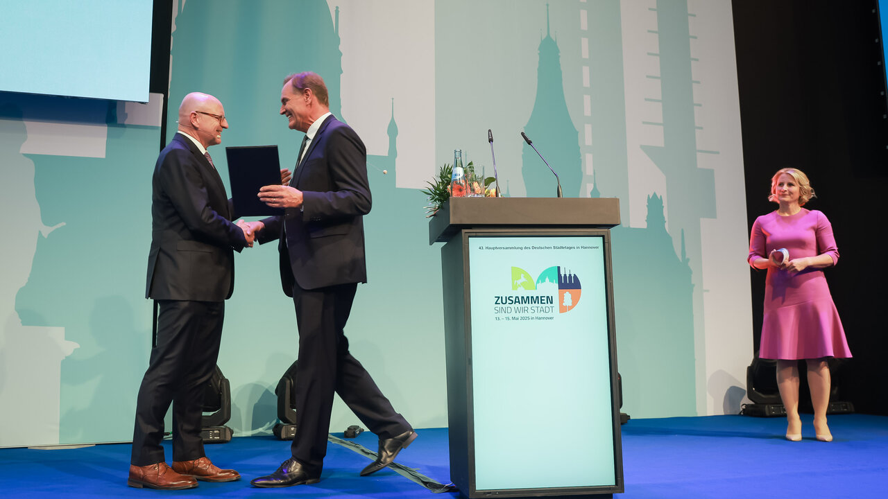 Burkhard Jung schüttelt Markus Lewe auf einer Bühne die Hand und überreicht ihm eine Ehrenmitgliedsurkunde, Moderatorin Susan Link steht im Hintergrund.