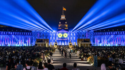 Das Bild zeigt die Schlosslichtspiele Karlsruhe und das entsprechend angeleuchtete Schloss im Hintergrund. Im Vordergrund ist die Vielzahl an Besuchern zu sehen.