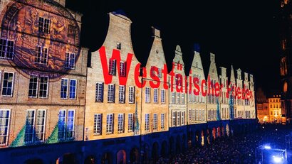 Das Foto zeigt eine Häuserreihe bei Nacht, die beleuchtet wird. Unter anderem sind eine Münze und der rote Schriftzug "Westfälischer Frieden" auf der Häuserwand zu erkennen.