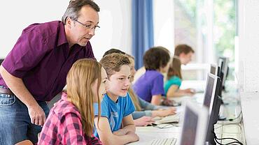 Digitale Schule: Schüler und Schülerinnen arbeiten an Computern, im Hintergrund der Lehrer.