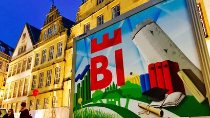 Auf dem Foto sieht man angeschnitten eine Fußgängerzone vor einer Häuserreihe. Vor der Häuserreihe hängt ein großes Plakat, das in etwa die Hälfte des Fotos einnimmt. Auf diesem erkennt man unter anderem eine Landschaft und unterschiedliche Gegenstände wie ein Buch, eine Schriftrolle und einen schiefen Turm. 