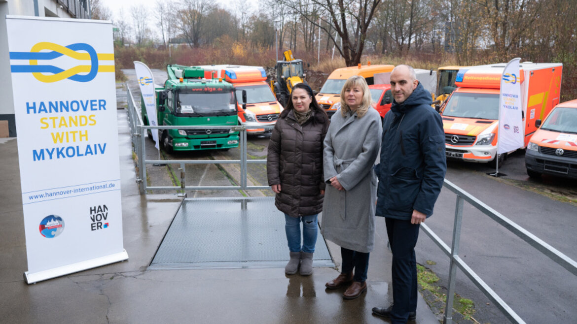 Drei Menschen stehen vor einer Reihe von Fahrzeugen und neben einem Banner mit der Aufschrift "Hannover stands with Mykolajiw"