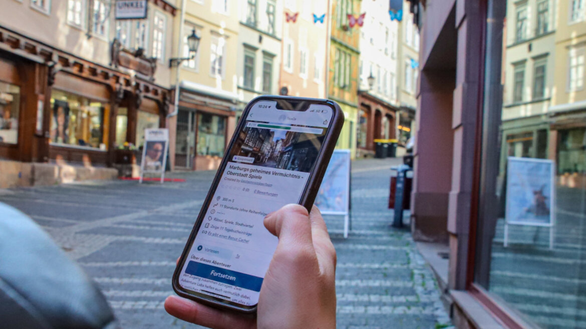 Eine Person steht in einer Straße in der Altstadt Marburgs und hält ein Smartphone in der Hand.