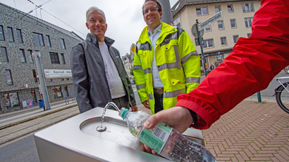 Am neuen Trinkwasserbrunnen am Konrad-Adenauer-Platz wird eine Flasche befüllt, im Hintergrund stehen zwei Personen.