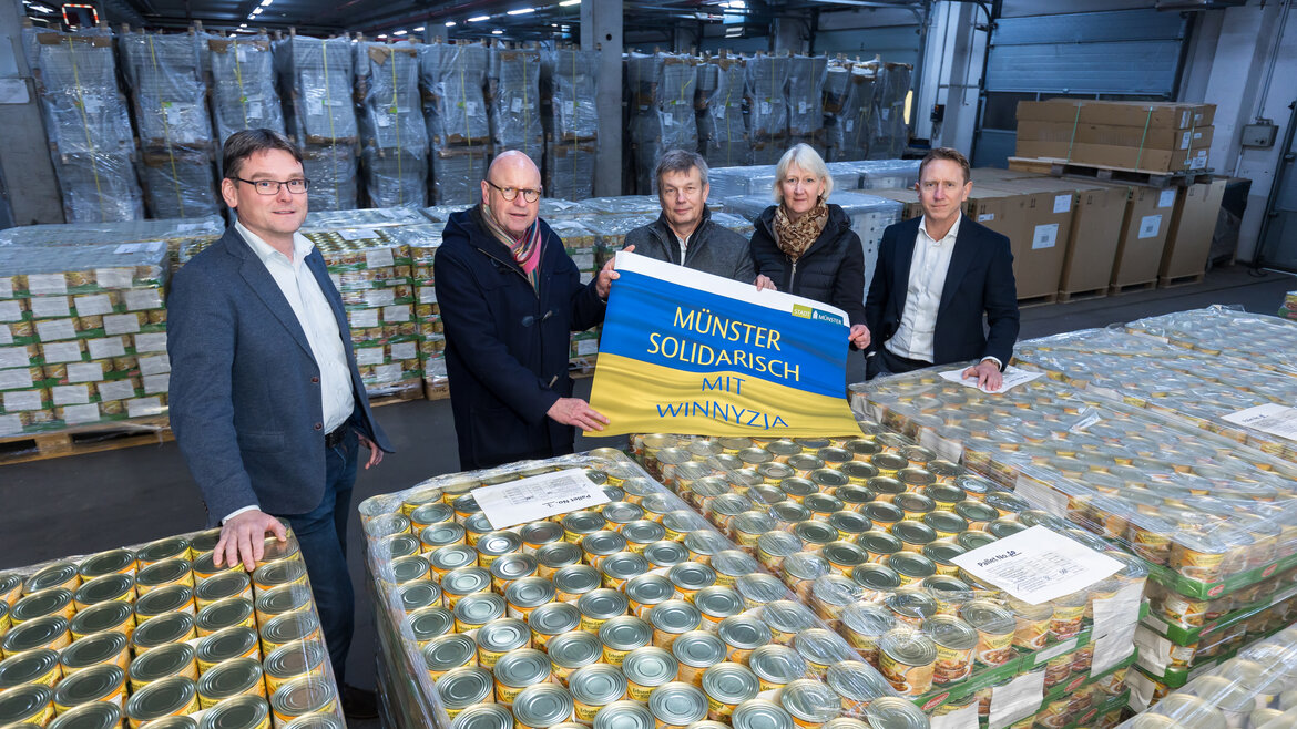 Eine Gruppe von Menschen steht vor Paletten mit Lebensmittelkonserven und hält eine ukrainische Flagge mit der Aufschrift "Münster solidarisch mit Winnyzja"