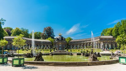 Auf dem Foto ist ein großes erhabenes Gebäude und davor ein große Wasseranlage mit Springbrunnen abgebildet: die Eremitage in Bayreuth.