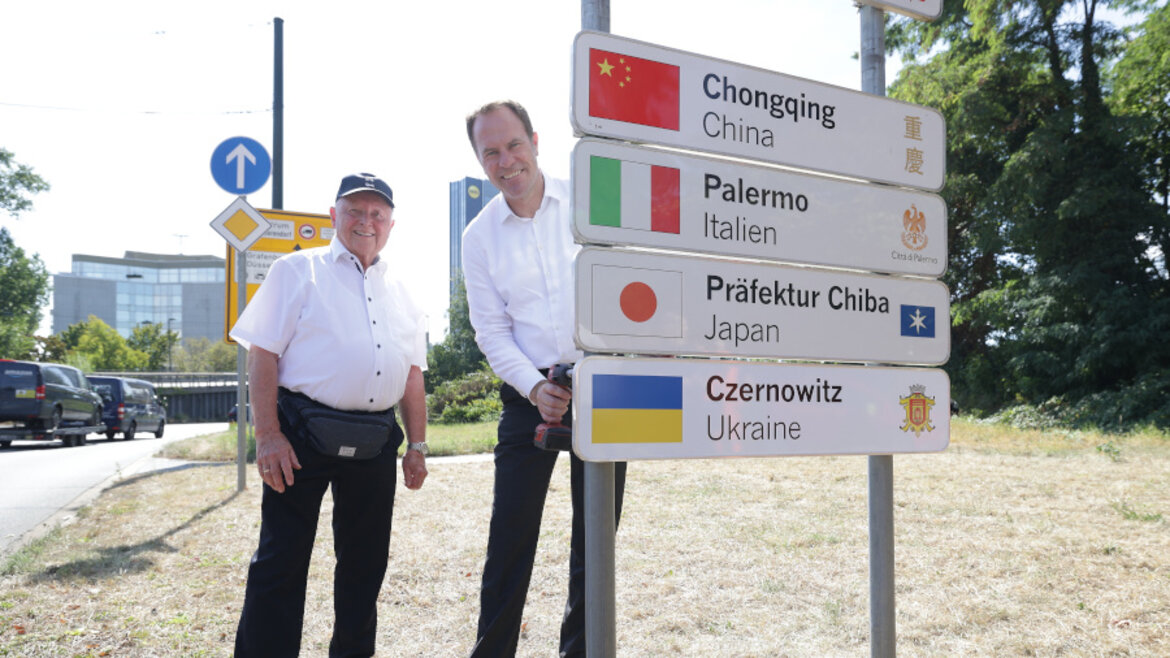 Zwei Männer vor Schildern mit Namen der Partnerstädte